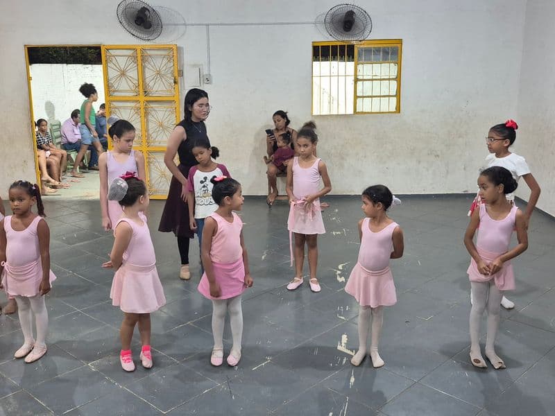 Crianças em uma sala de aula participam da oficina de ballet do projeto Realização. Várias alunas vestem collant e saia rosa, enquanto outras usam roupas casuais. Uma professora, de blusa preta e saia longa escura, está no centro orientando o grupo espalhado pelo salão.