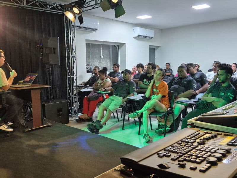 Imagem da Sala de aula com o professor falando ao microfone e os alunos sentados em carteiras ouvem atentamente.