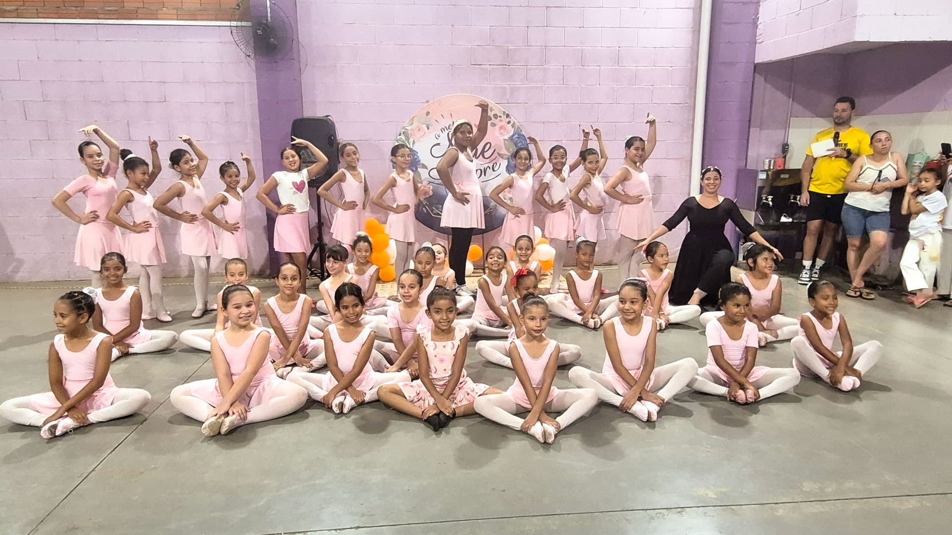 Grupo de meninas alunas da oficina de ballet em uma pose para foto vestidas com collants e sapatilhas, a frente meninas sentadas em pose de borboleta e atrás em pé meninas em pose de ballet, a direita do grupo a instrutora vestindo roupas pretas.